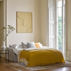 A styled bedroom interior with a bed dressed in white bedding and a mustard yellow textured throw, set against cream walls with a framed abstract print and a tall window.