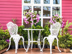 White metal chairs and table in front of a pink building with greenery and flowers.