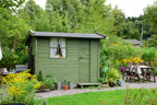 Green garden shed in a lush garden with trees and flowers.