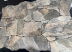 A stone-look decorative wall panel or tile sample with irregular faux-stone shapes in mixed gray, beige, brown, and charcoal tones, photographed against a black background.