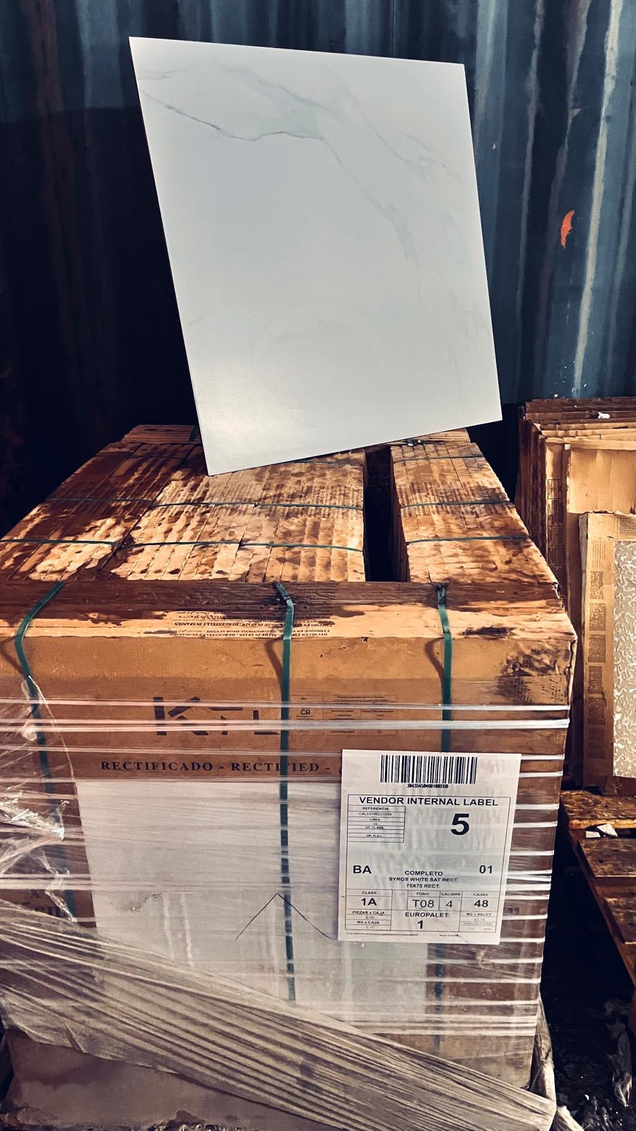 A large white rectangular tile with subtle gray veining stands upright on top of stacked wooden pallets wrapped in clear plastic. A shipping label is visible on the pallet, and the background shows a dark corrugated container.