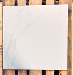 A square white marble-look tile with soft gray veining, displayed flat on a rustic wooden slatted surface.