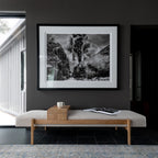 A large black and white framed art print depicting a steam train in a mountain landscape, displayed on a dark gray wall above a light wood bench in a modern interior setting.