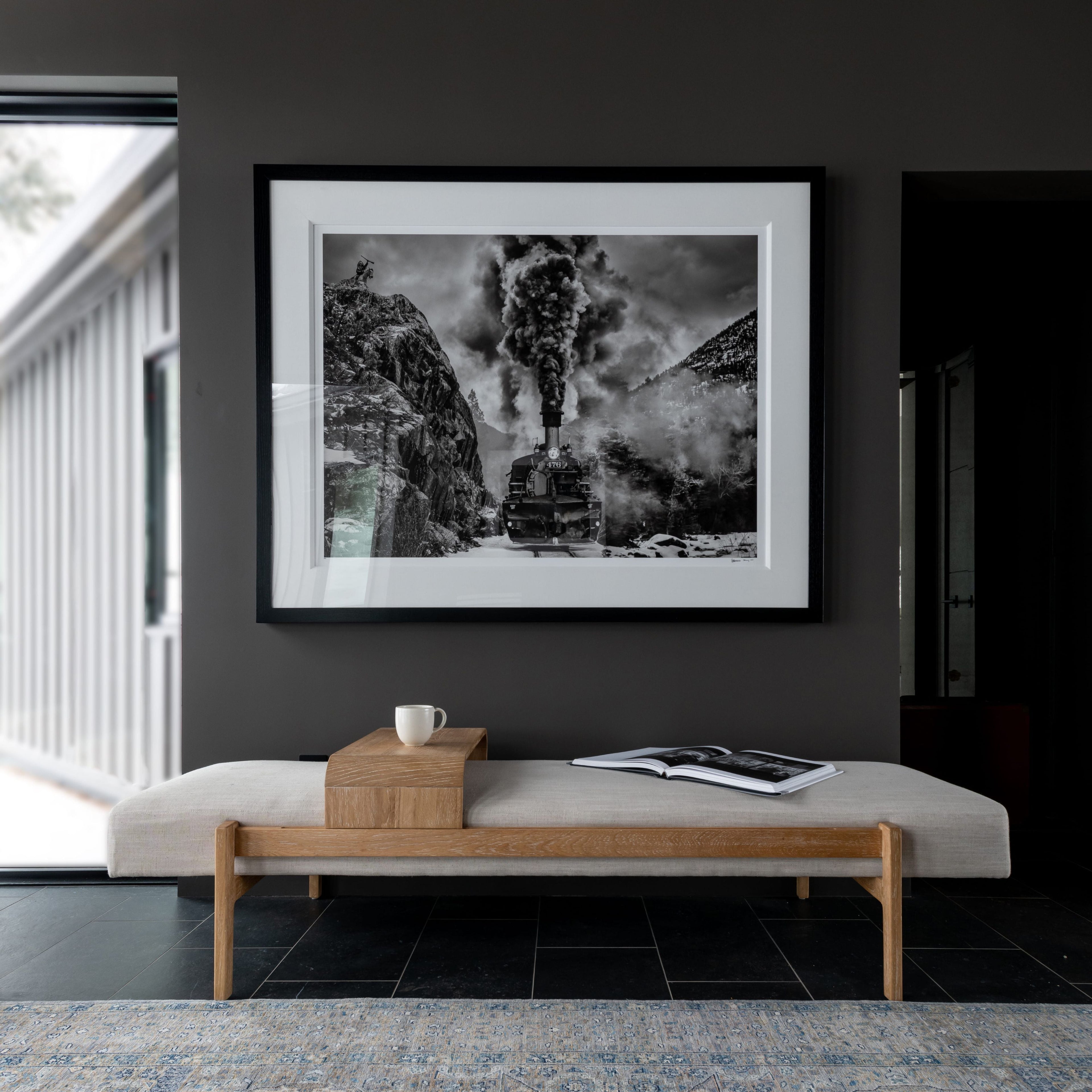 A large black and white framed art print depicting a steam train in a mountain landscape, displayed on a dark gray wall above a light wood bench in a modern interior setting.